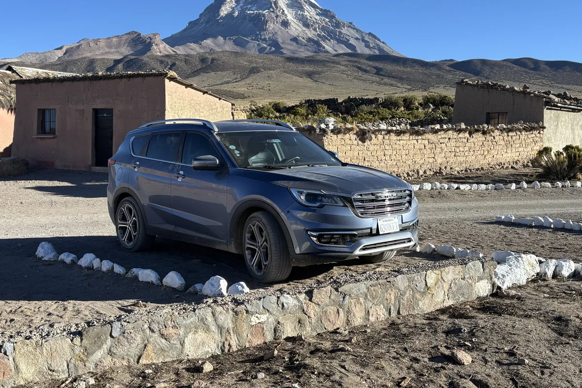 Fat Cat’s Jetour SUV 2025, blue-grey, parked by adobe houses with the snow-capped Sajama volcano behind